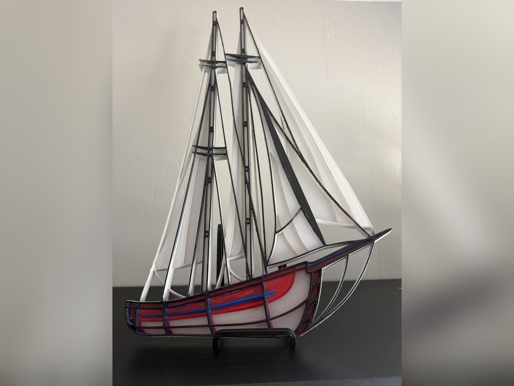A stained-glass style sailboat with white sails and a red and blue hull, standing on a small stand